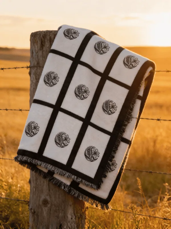 Nine panel sublimation blanket with black grid border and botanical medallion print folded over a weathered wooden fence post in a golden Saskatchewan prairie field at sunset