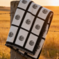 Nine panel sublimation blanket with black grid border and botanical medallion print folded over a weathered wooden fence post in a golden Saskatchewan prairie field at sunset