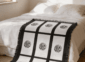 Nine panel sublimation blanket with black grid border and botanical medallion print layered at the foot of a bed with white linen bedding in a neutral boho bedroom