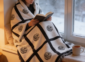 erson wrapped in a nine panel sublimation blanket with black grid border sitting in a cosy window seat reading with a mug of tea and snow falling outside