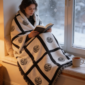 erson wrapped in a nine panel sublimation blanket with black grid border sitting in a cosy window seat reading with a mug of tea and snow falling outside