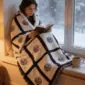 erson wrapped in a nine panel sublimation blanket with black grid border sitting in a cosy window seat reading with a mug of tea and snow falling outside