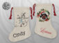 wo natural linen Christmas stockings with red drawstring ties — one with an ice skating snowman and the name Cindy in black, the other with a watercolour dachshund in a Santa hat wreath and the name Loriann in red script